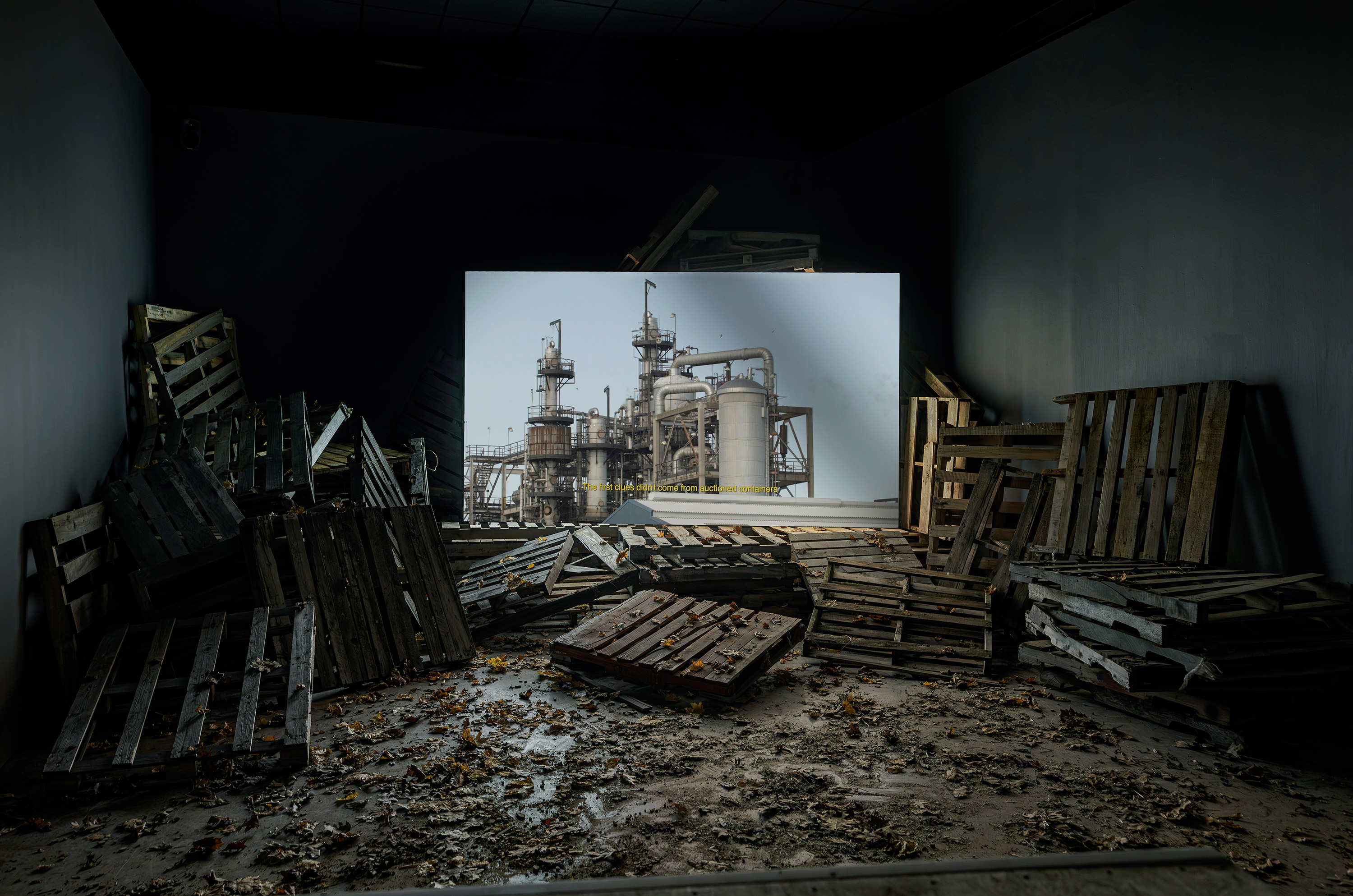 Photo of a projection of an industrial factory in the gallery space surrounded by wooden pallets stacked against each other on a floor of dried leaves