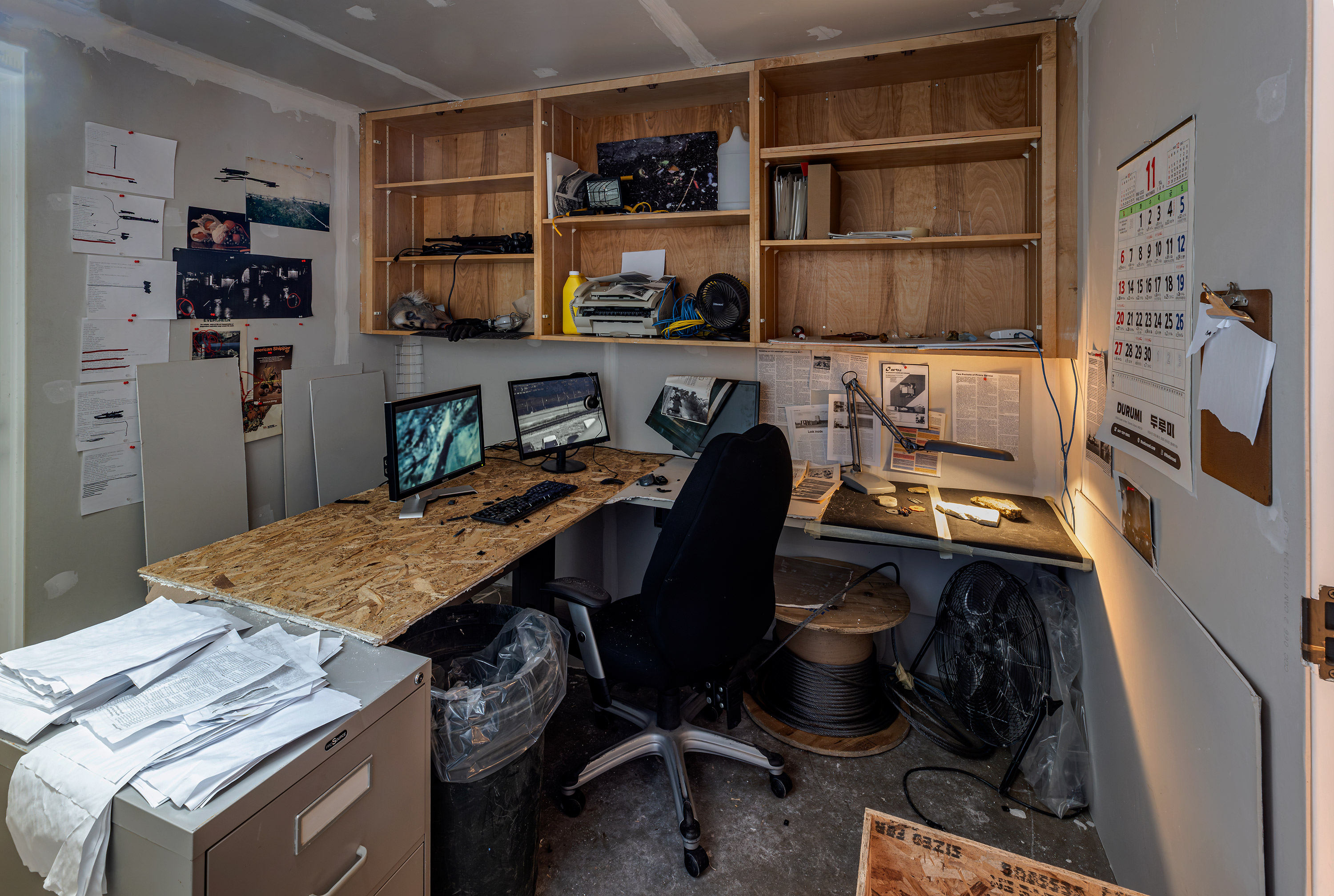 Photo of the inside of the Conspiracist "C's" office space filled with scattered papers, a wooden desk with computer screens and assorted photos on the walls