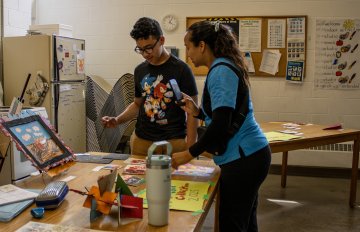 Young person standing beside a display of their artwork, a parent leans over the table to look at the drawings and illustrations