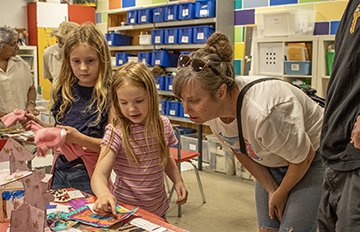 Two children standing in the KWAG art studio beside a table of their artwork with a parent crouching over to closely examine artwork 