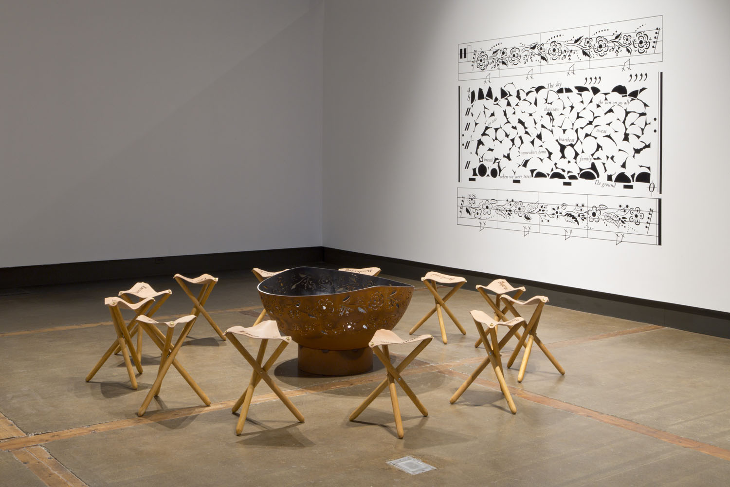 Installation view of Surrounded/Surrounding, a circular arrangement of leather stools around a copper fire bowl etched with a floral pattern, in front of a white gallery wall with a black vinyl transfer of a large pattern of shapes and text