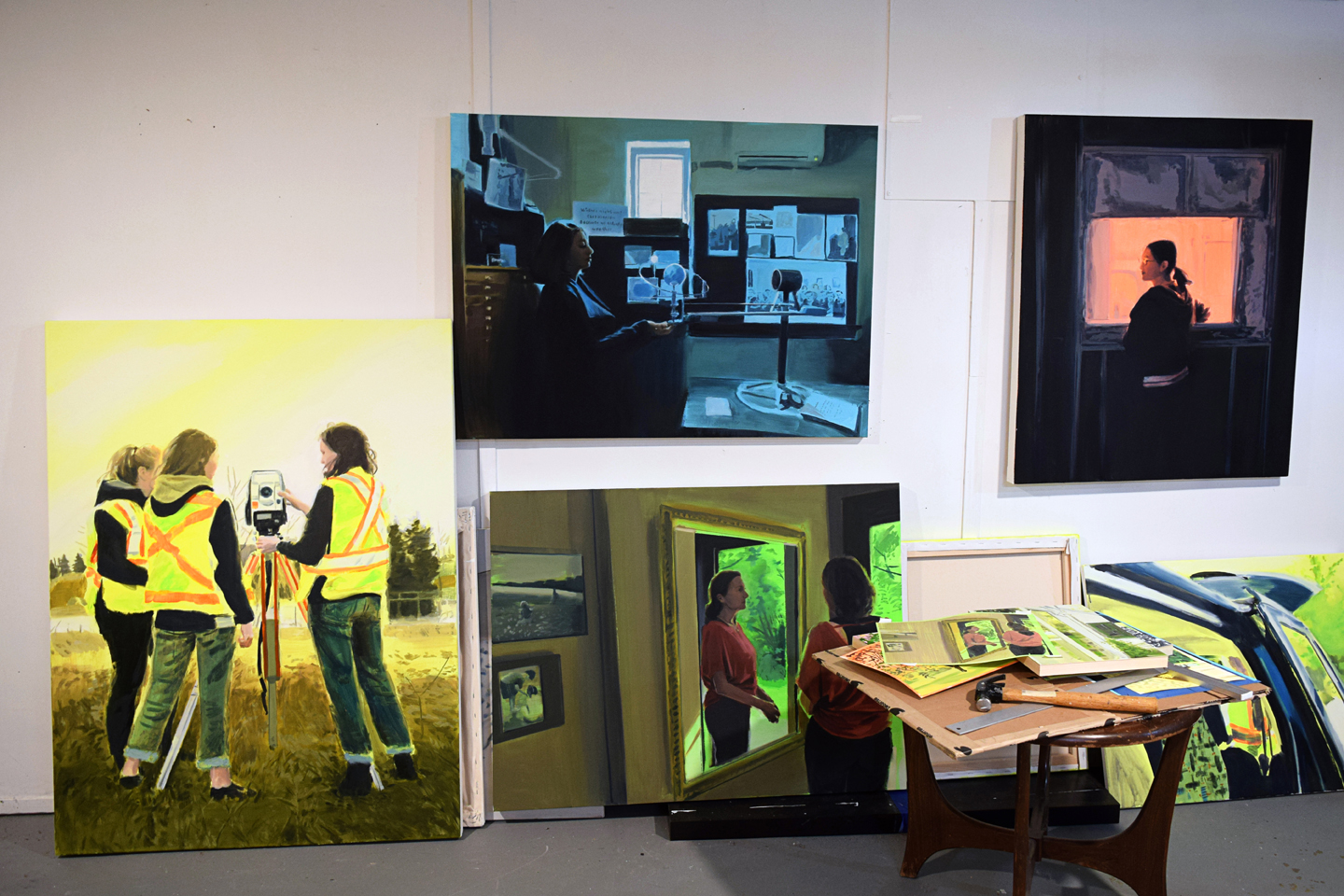 View of Amanda Rhodenizer's studio wall showing four paintings in progress of working women in saturated colour settings; a small studio table sits in the foreground