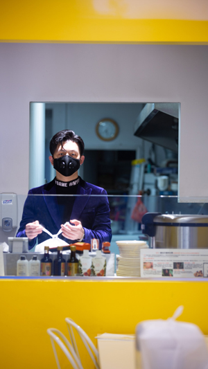 Vertical still image of a take-out restaurant worker in a facemask standing behind a glass counter while tying a white plastic bag