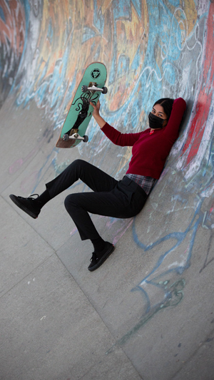 Vertical still image of a female skateboarder in dark clothing and a black facemask holding up her green skateboard while reclined against the side of a graffiti-covered half-pipe