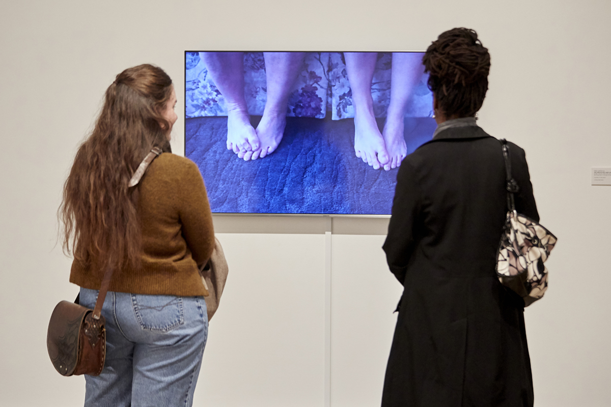 Two women stand before Erika DeFreitas' video work of two pairs of feet shown on a wall-mounted screen in the Gallery