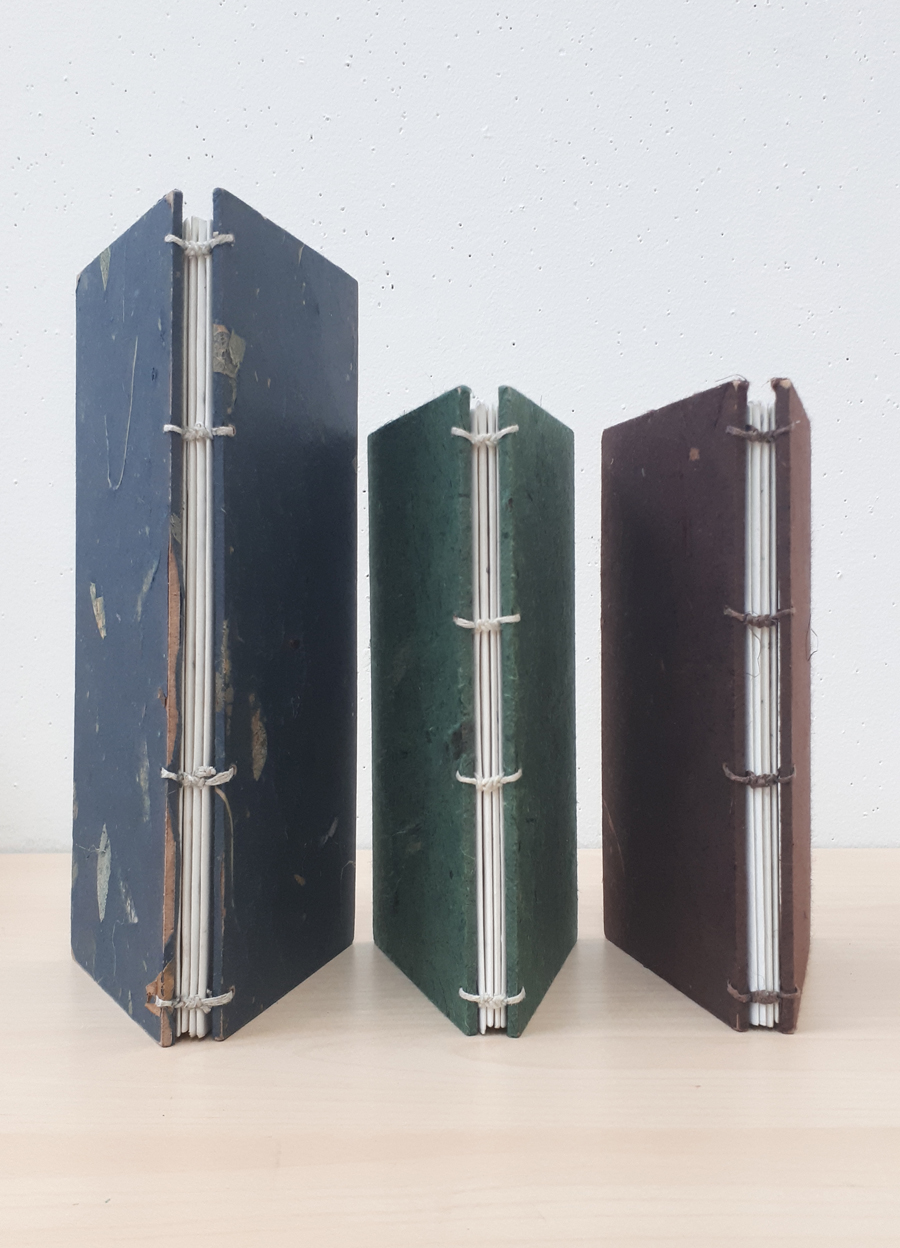 Three handmade sketchbooks bound with blue, green and maroon covers sit upright with their spines facing the viewer