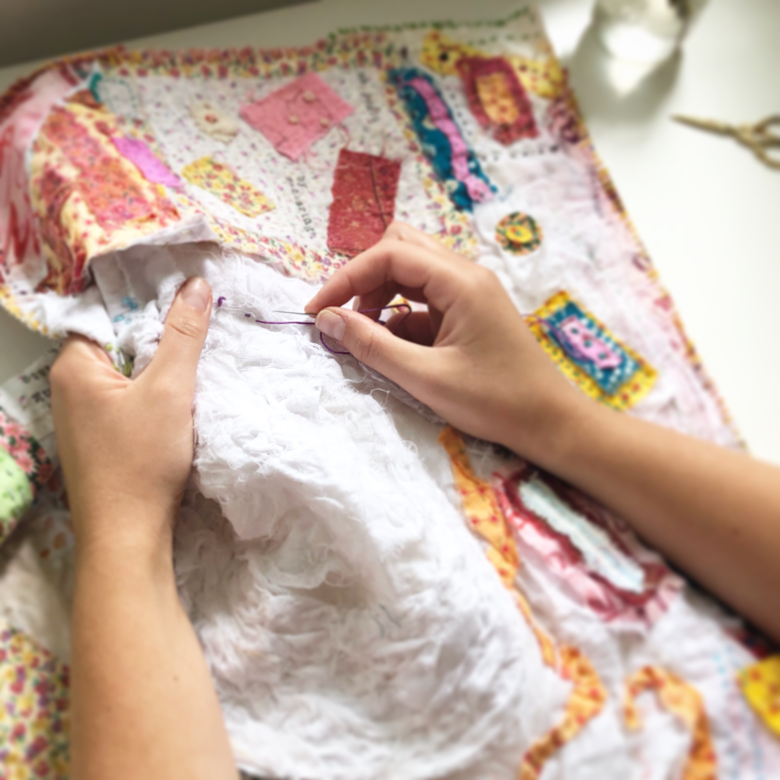 A pair of female hands holding a large piece of white fabric partially covered in pastel-toned embroidery and patches while stitching with dark red thread