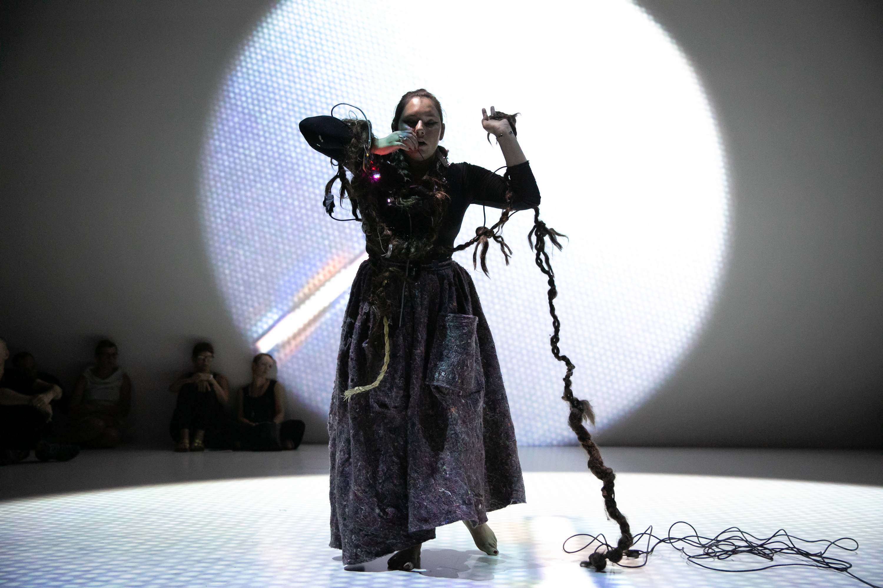 Kite performing in a long dark dress standing on a white-lit circle in a darkened space; a long knotted braid woven with wires extends from her head to the floor