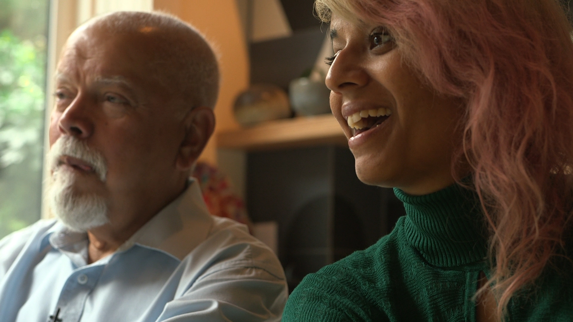 A fillm still from Arrival Archives shows a ponderous older man with a white goatee and a younger laughing woman with pink-dyed hair in a warm domestic interior space