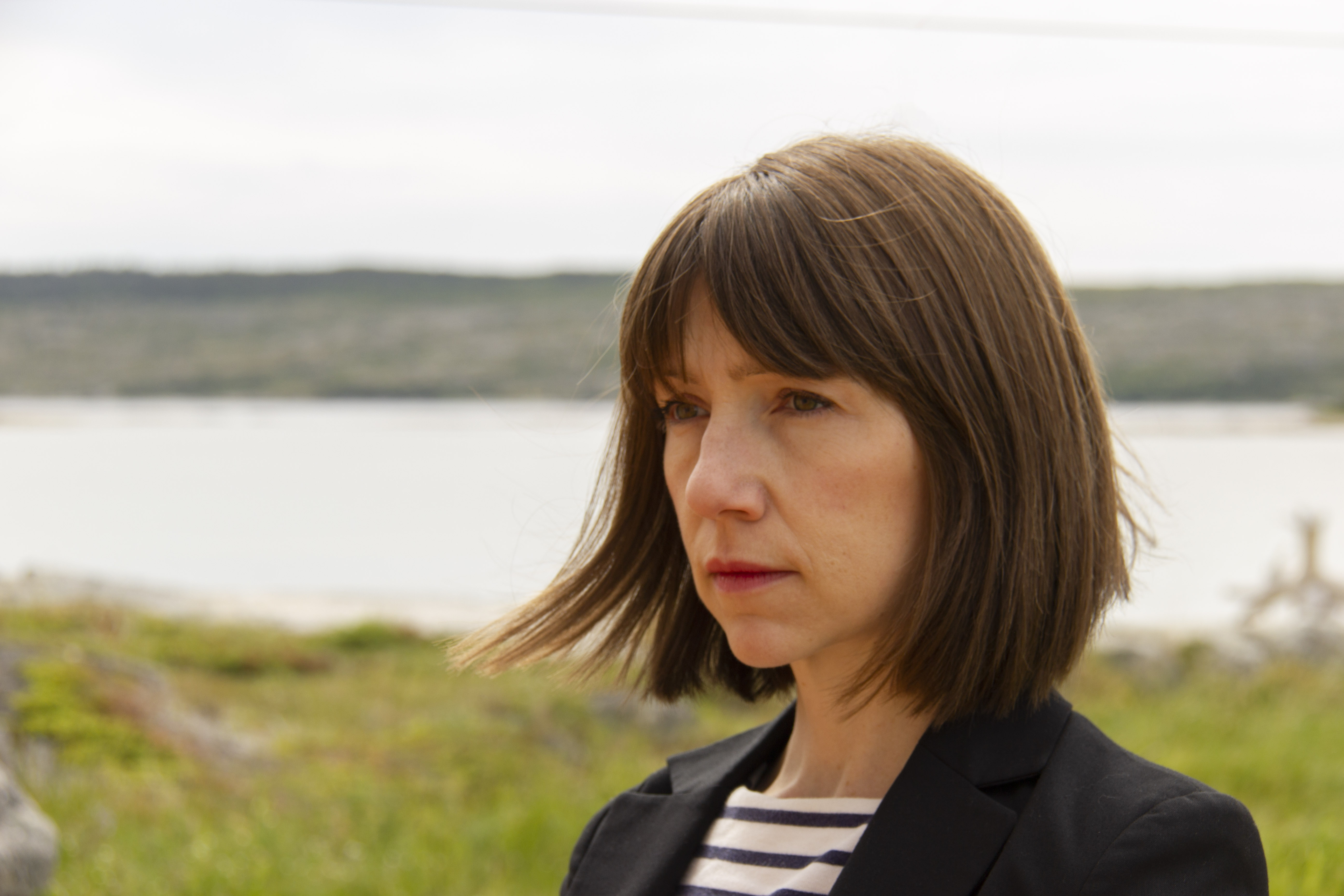 Photo of Alexandra McIntosh, a woman with shoulder-length brown hair gazing off-camera against a background of grass and water