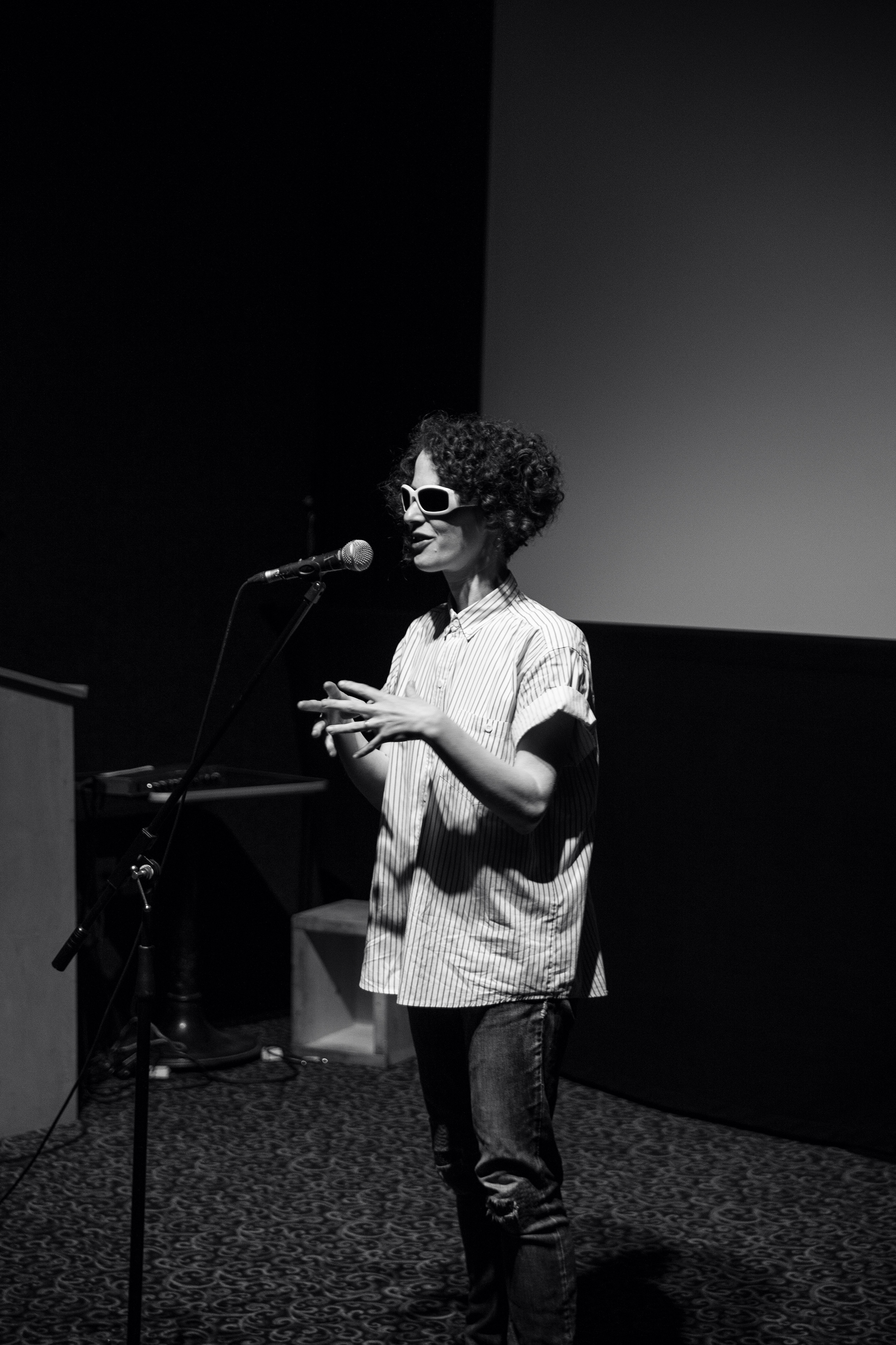 Black and white photo of Aislinn Thomas, a woman with short curly dark hair wearing dark glasses and a white short-sleeved shirt speaking at a microphone in a dark presentation space