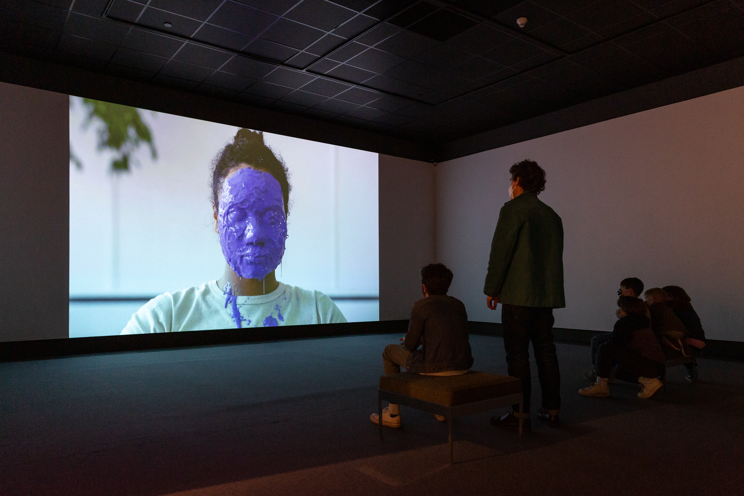 A small group of gallery visitors standing, seated and crouched on a grey carpeted area in front of a large video projection, filling the wall, of a woman in a white t-shirt with purple casting latex covering her face and spattering her clothes