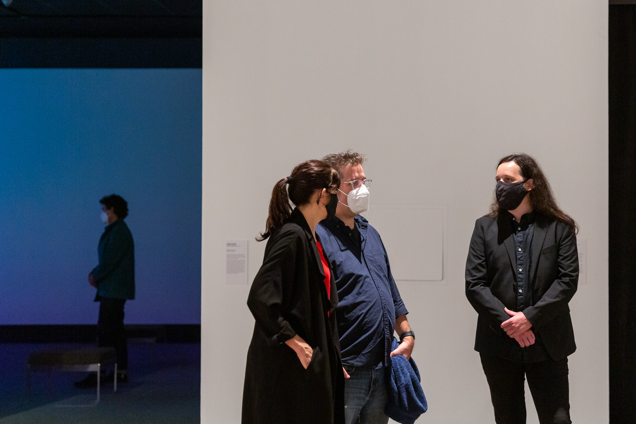 Two gallery visitors stand near Darryn Doull, a man in a dark suit and matching face mask, in the gallery in front of a white wall; a fourth person is visible in a blue-lit room behind them