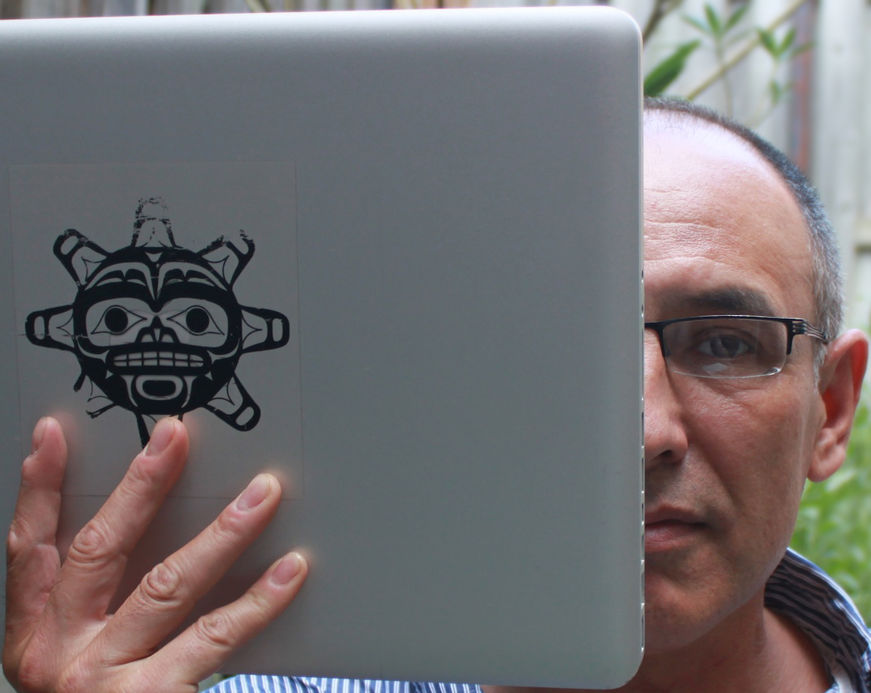 Archer Pechawis, an older Cree man with close-cropped grey hair, holds up a silver laptop to cover half his face