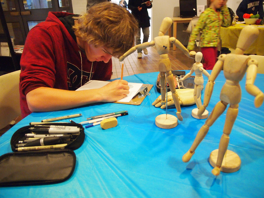 A teenage boy in a red hoodie seated at a table drawing intently from a series of wood mannekin figures