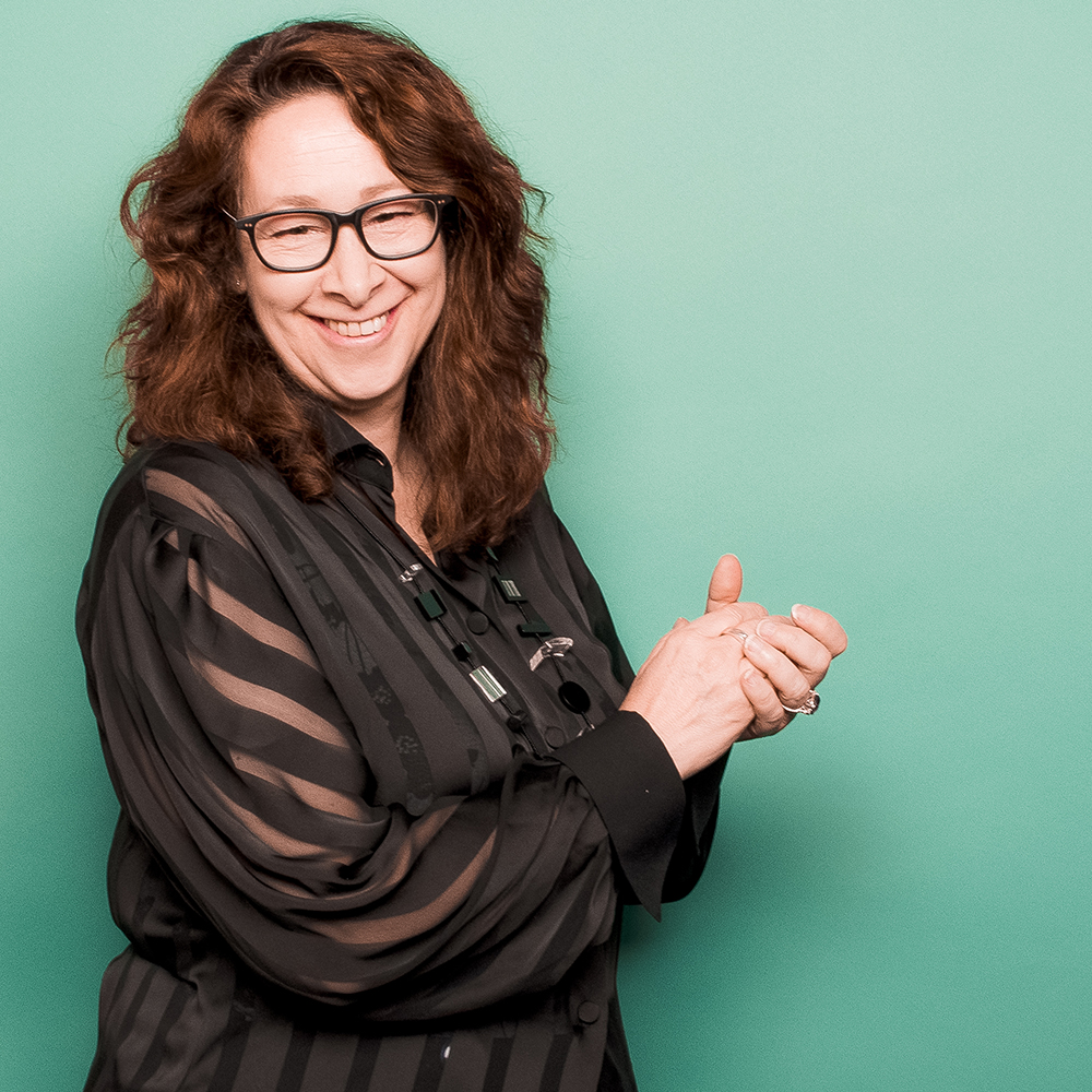 Photo of Caroline Robbie, a smiling middle-aged woman with light brown hair and glasses, posing against a mint green background