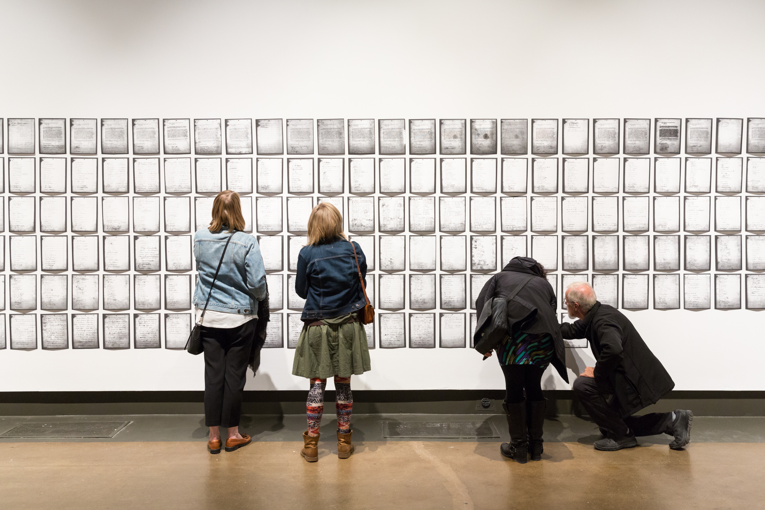 Photo of four visitors seen from behind as they examine Deanna Bowen's petition installation