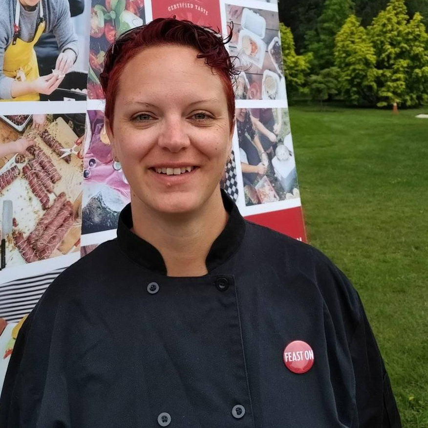Photo of Chef Stephanie Soulis, a smiling woman with close-cropped red hair wearing a black chef's jacket, standing outside in front of a photographic banner