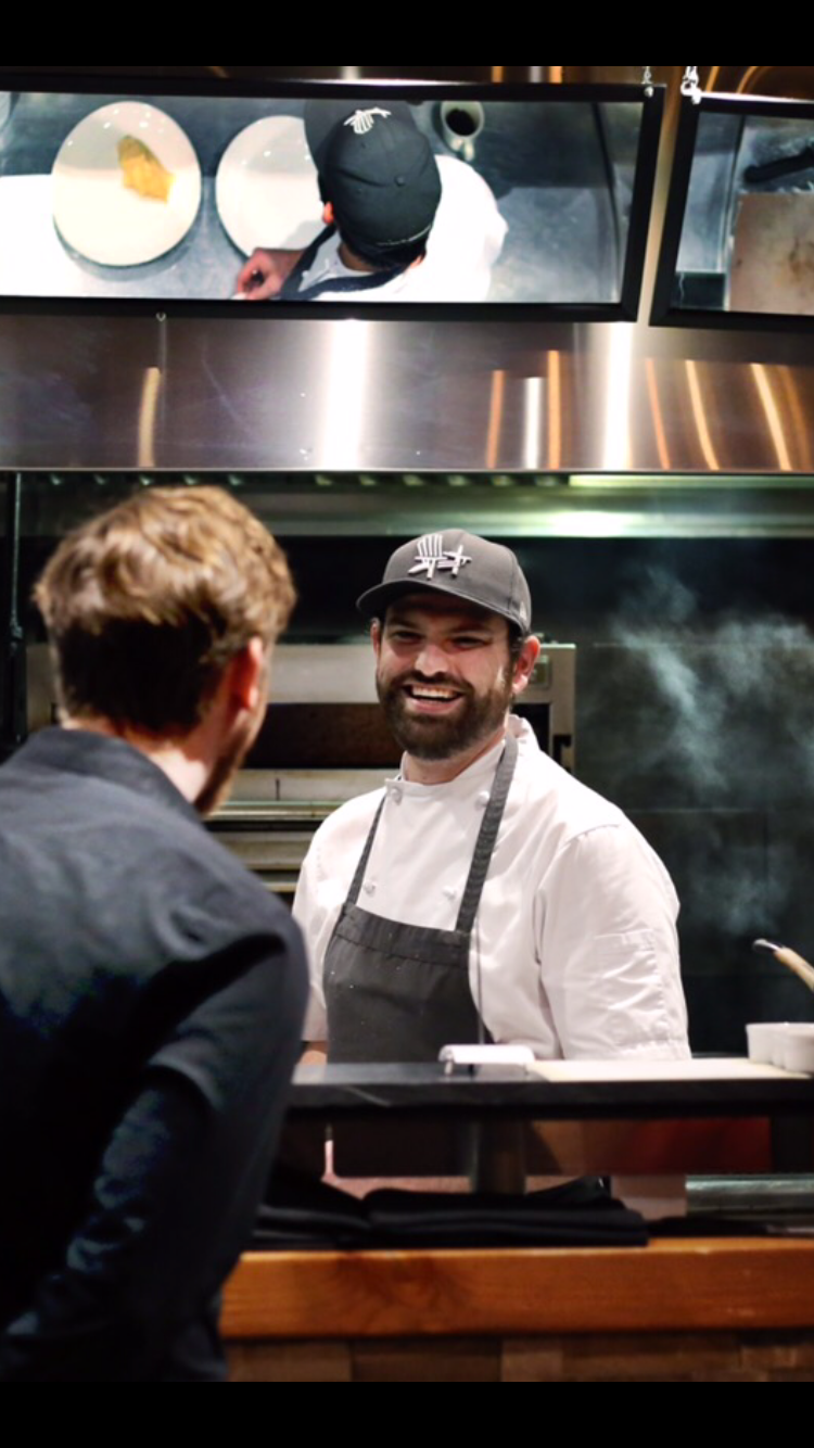 Photo of Chef Kyle Rennie, a young man laughing at the pass in a busy kitchen