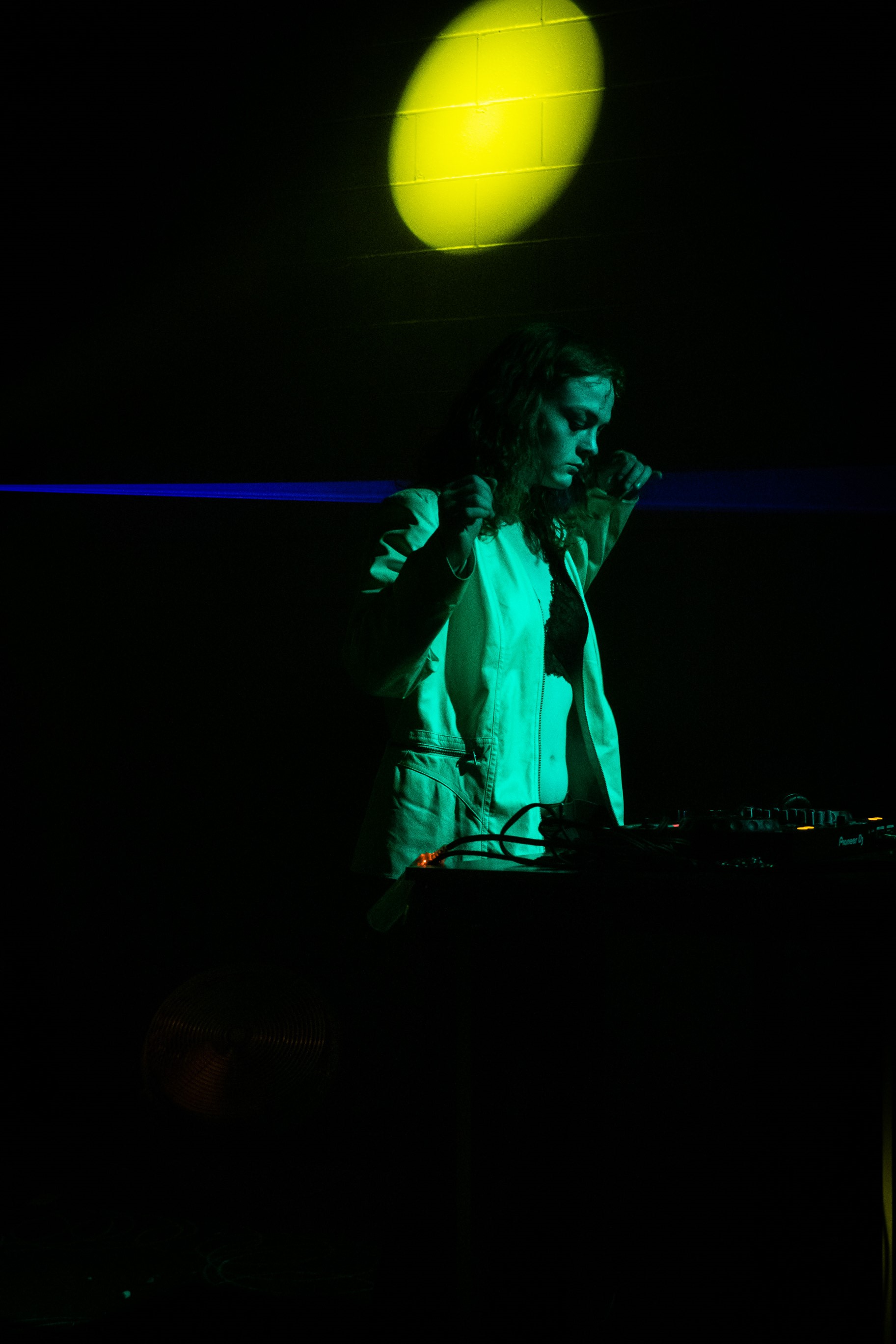 Photo of DJ Slayla, a green-lit woman isolated in a black space with a bright yellow circular light projected above her