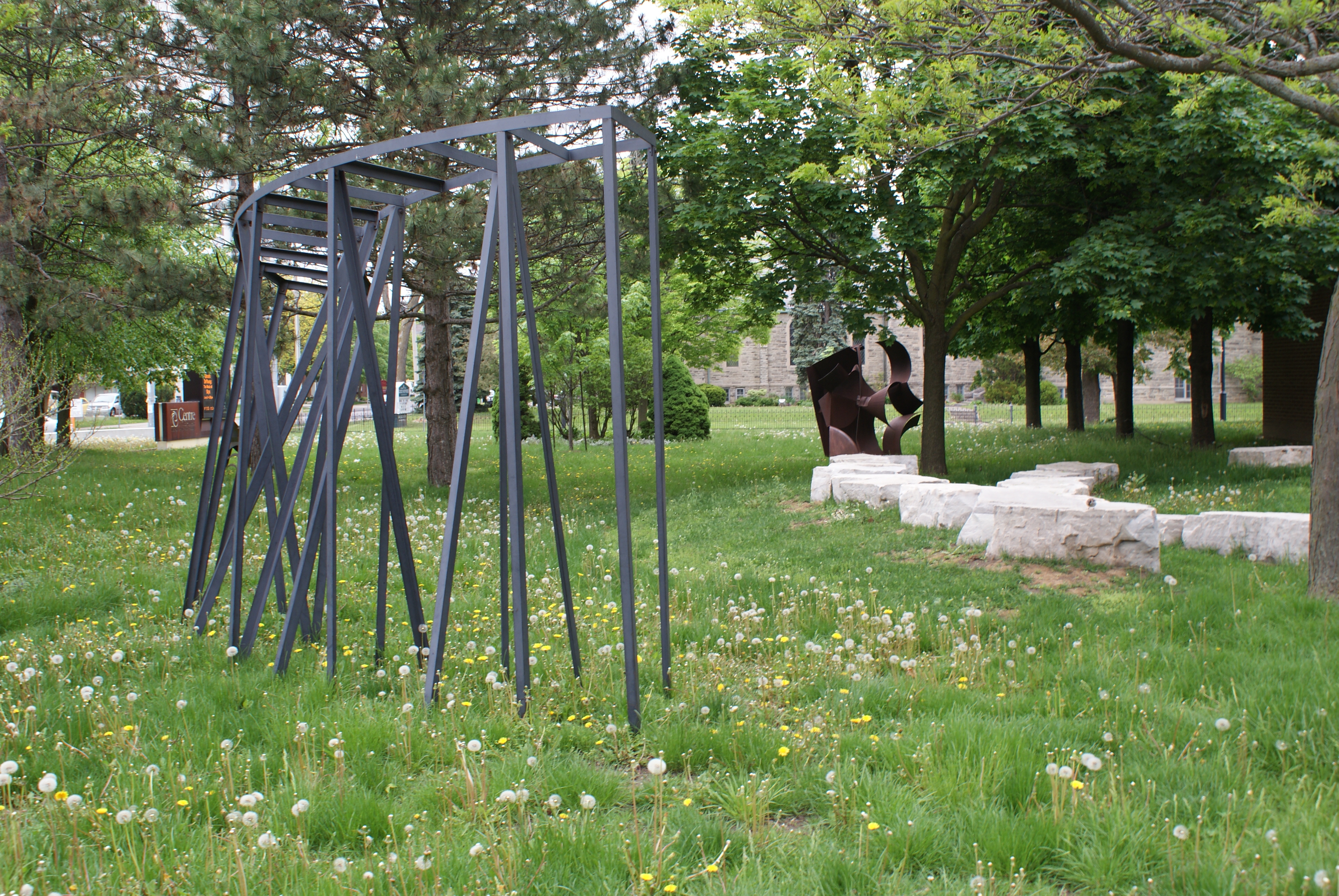 Photo of KWAG's sculpture garden featuring a large angular steel sculpture in the foreground of a grassy, tree-covered space
