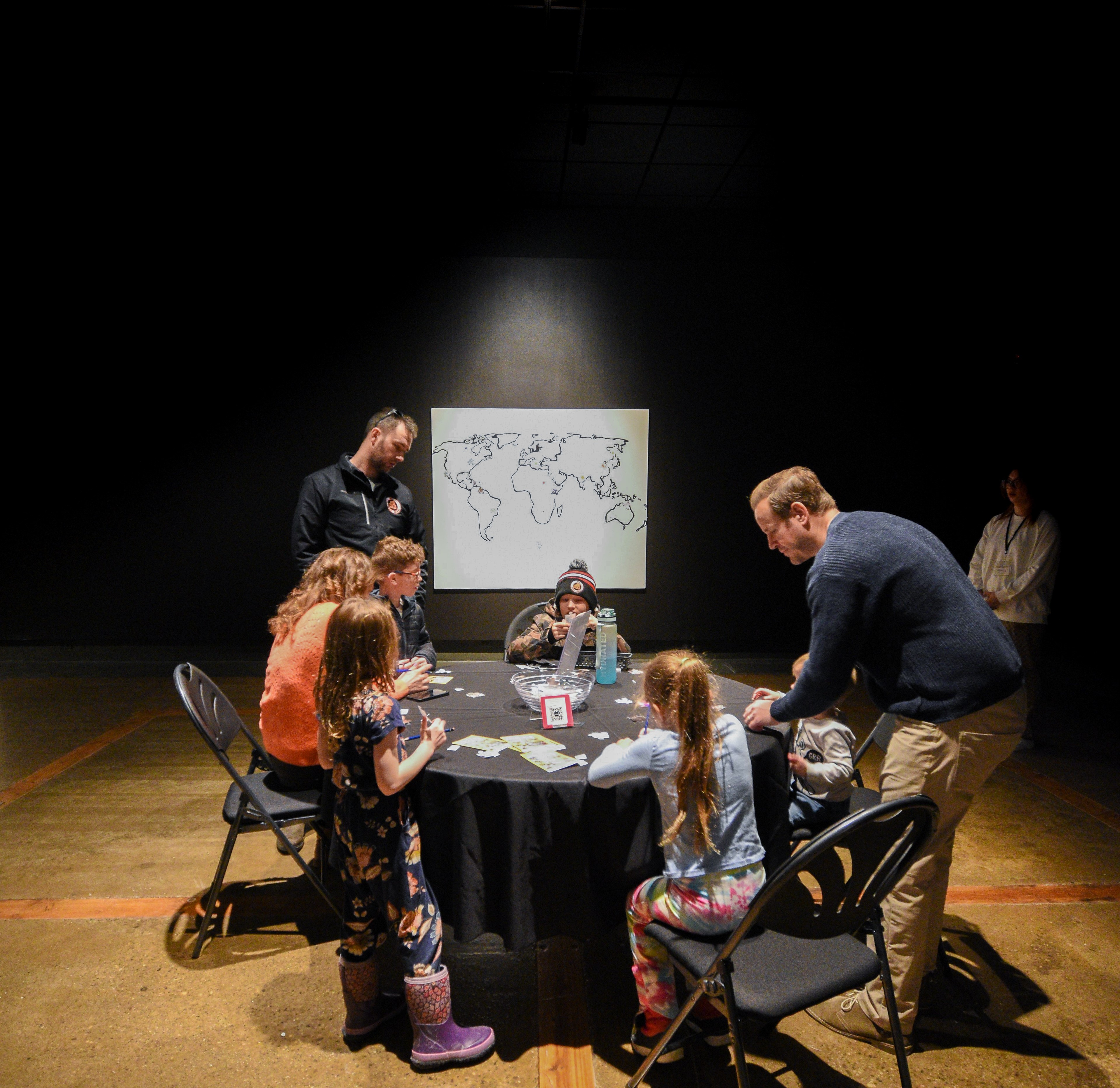A family sitting around a table in a black room with a outline of the world map on the wall.