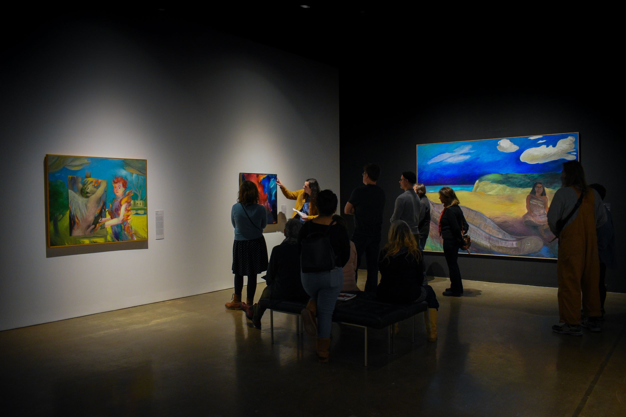 A group of gallery visitors in a dimly lit room listening to the curator speak. Looking at colourful paintings