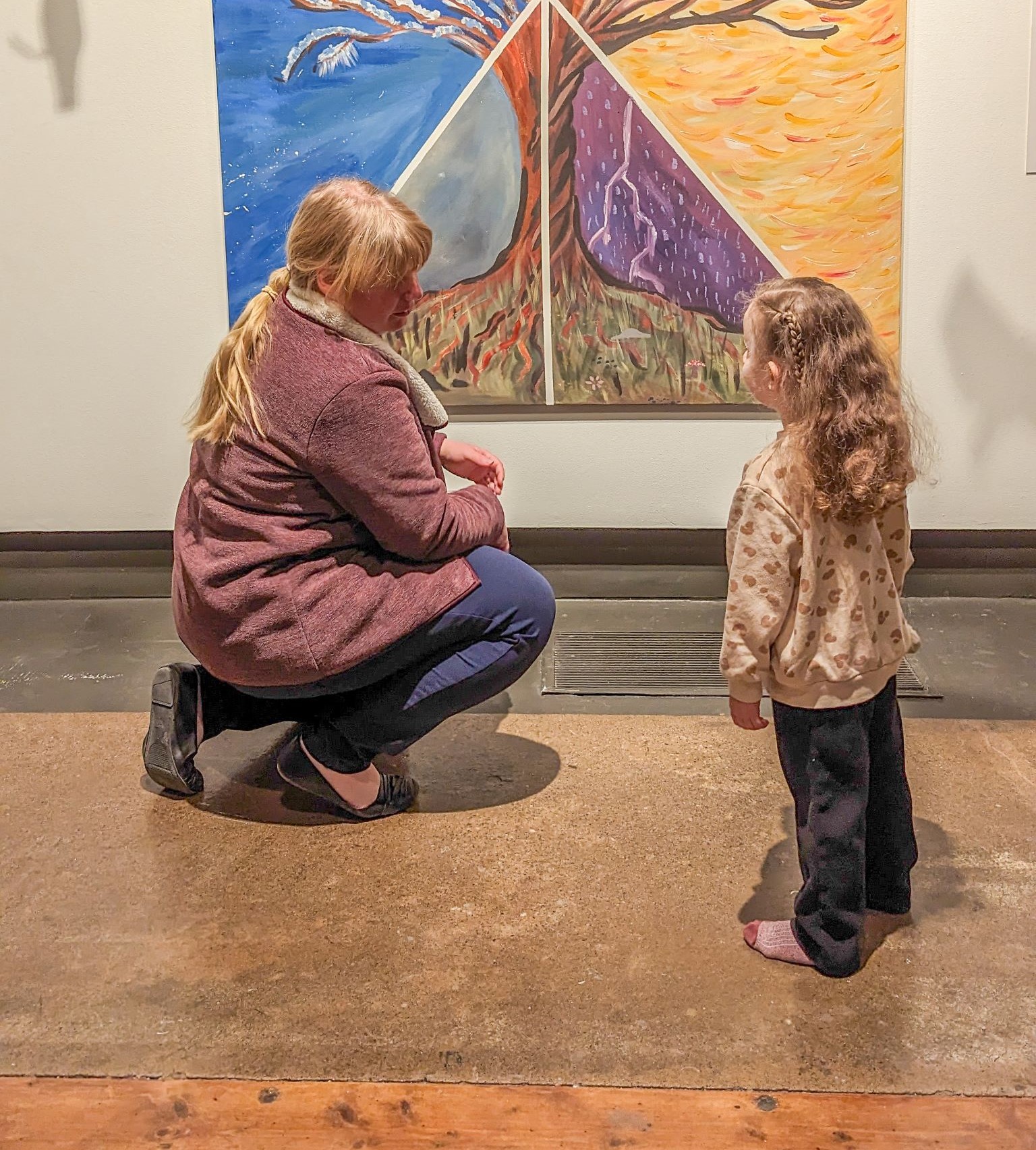 woman crouched beside a young girl looking at a larger painting of a tree