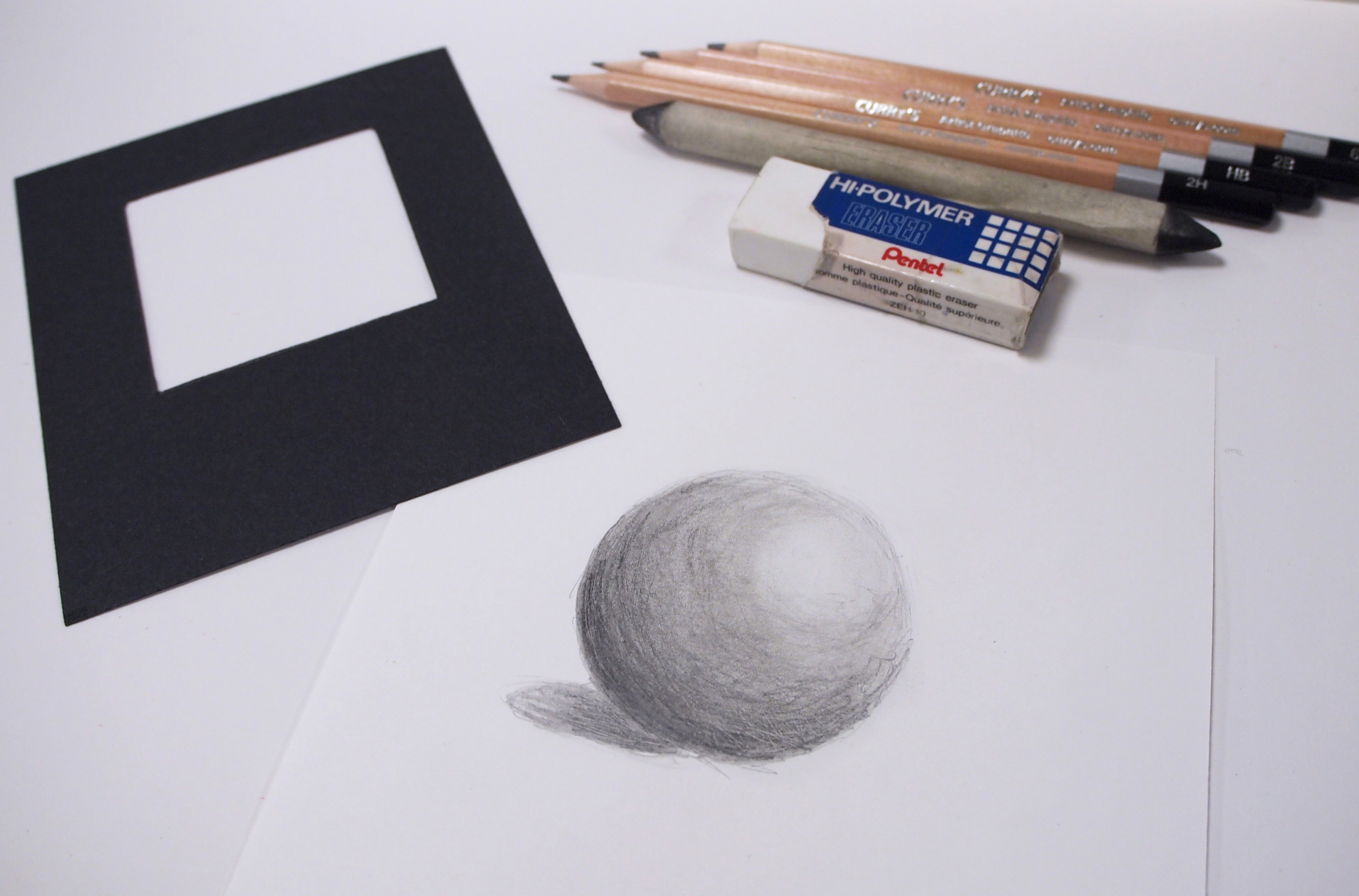 Photo of pencils, eraser, blending stomp and black viewfinder laying alongside a shaded drawing of a sphere