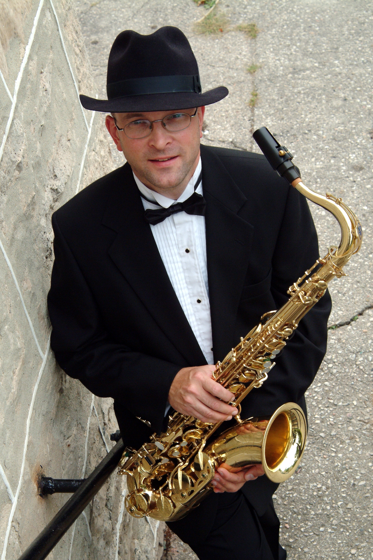 Photo of saxophonist Ernie Kalwa in a black tuxedo and black fedora, holding his saxophone