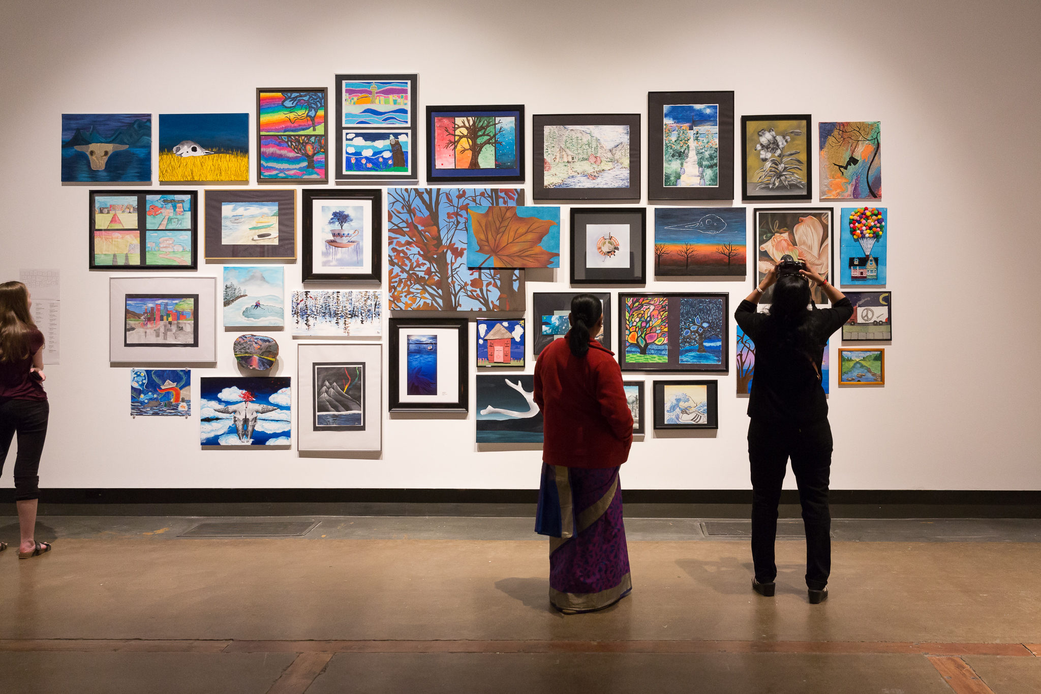 Gallery visitors seen from behind as they look at a gallery wall filled with a large array of framed student paintings and drawings