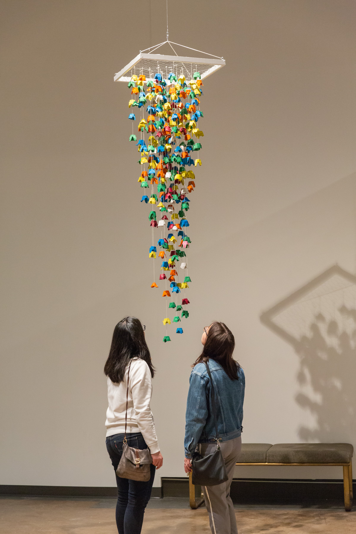 Two young women look up at a suspended sculpture of colourful shapes dangling down from a white pyramid frame in Expressions 44