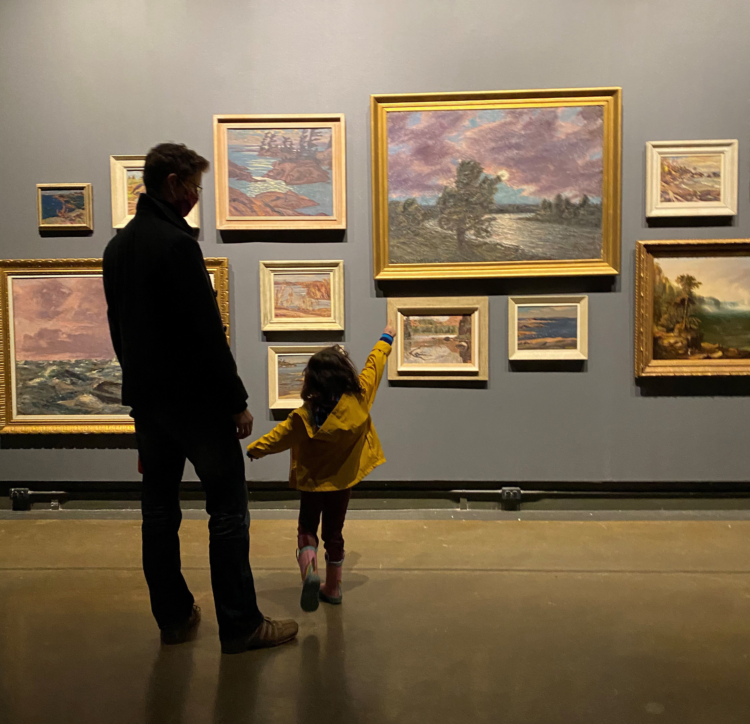 A man accompanies a small child in a raincoat and boots who points excitedly at a dark gallery wall filled salon-style with Canadian landcape paintings of various sizes