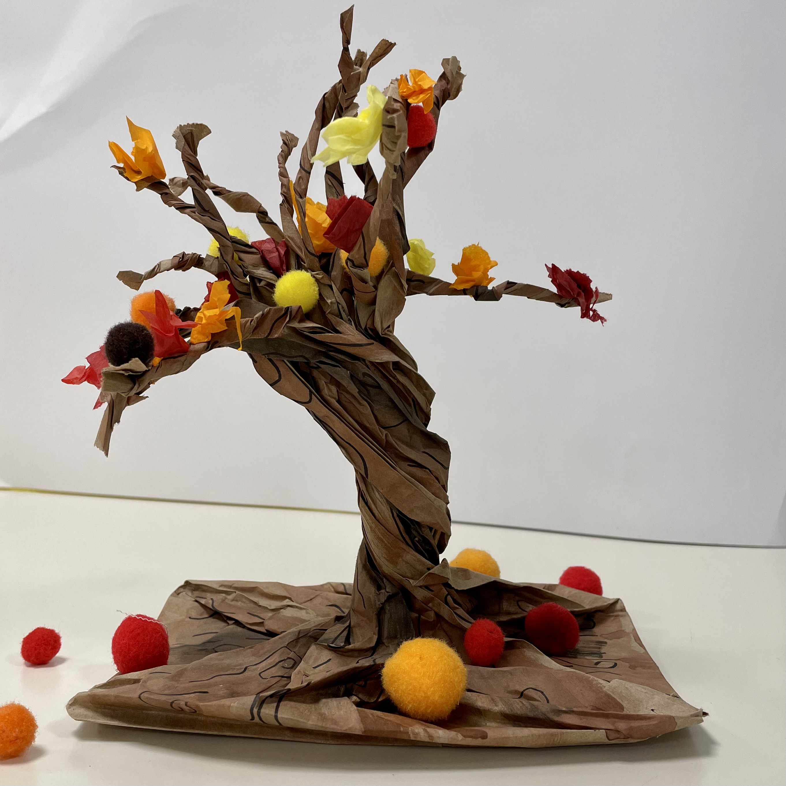 A brown paper tree decorated with red, orange and yellow tissue paper leaves and pom-poms displayed on a white surface against a white wall