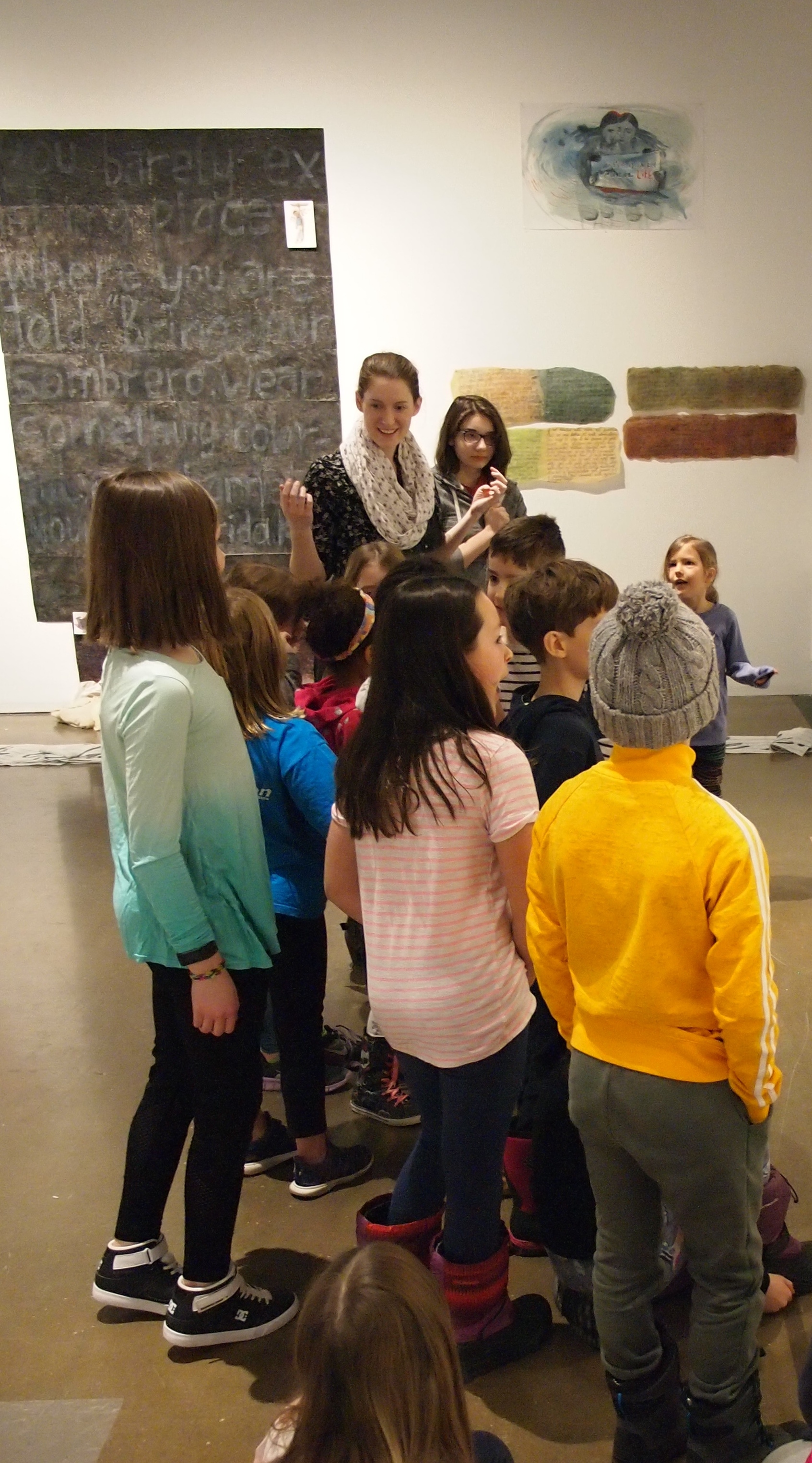 A group of school children stand together listening to a young woman in a dark top and light scarf explain artworks in a Lucie Chan exhibition of various drawings