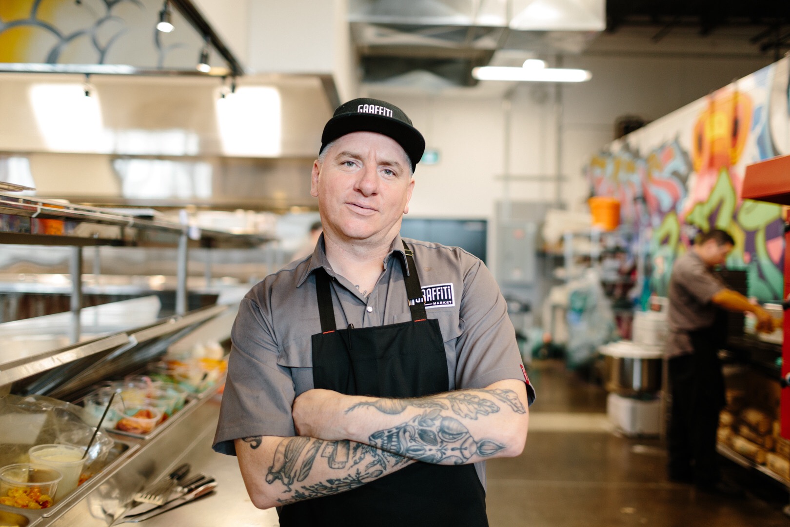 Photo of Chef Brian McCourt standing with tattooed arms crossed in the middle of a large restaurant kitchen