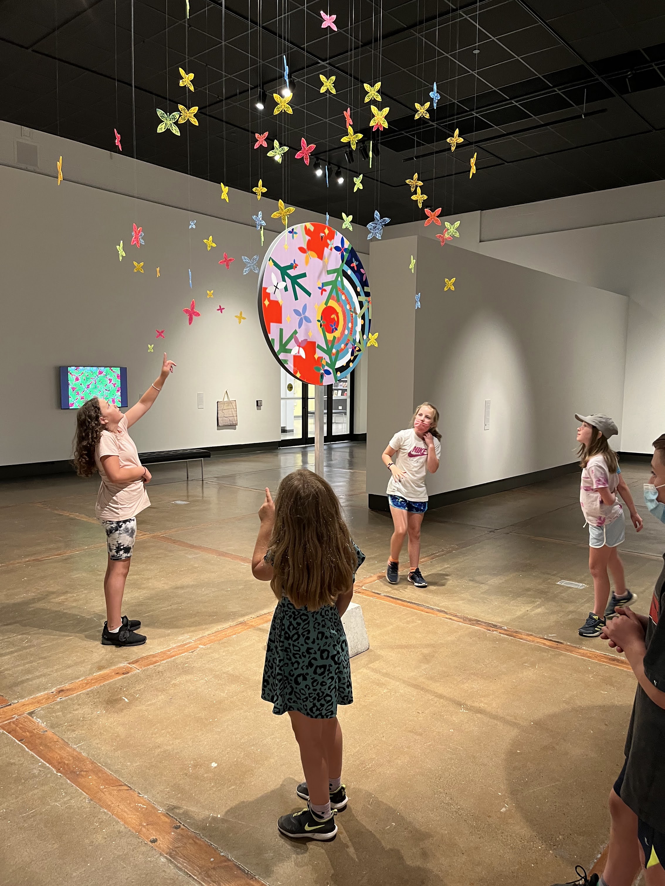 Four children stand in a circle around Jordan Bennett's artwork, one reaching up towards its suspended plexiglass butterflies