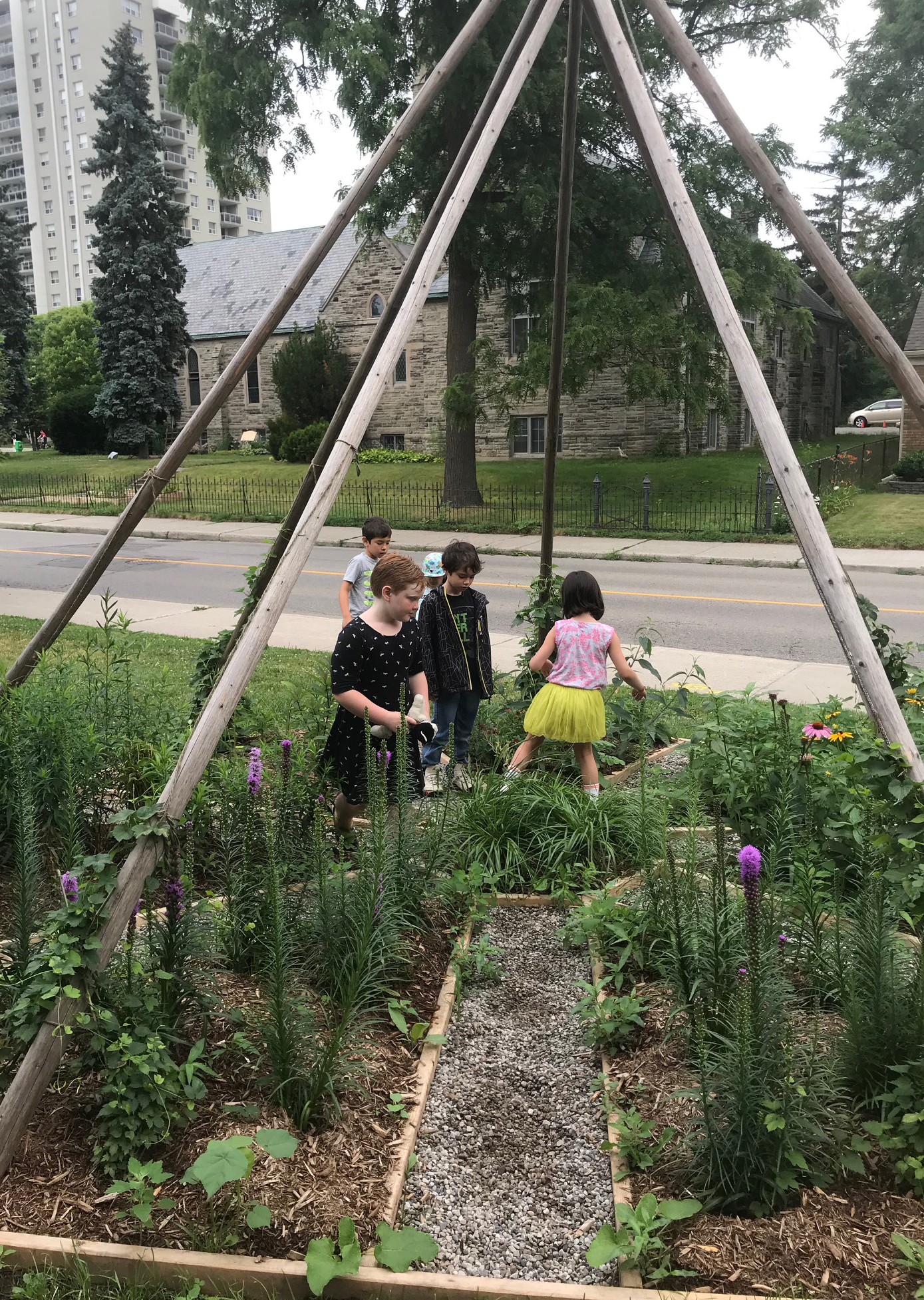 A small group of children explore the centre of a seven-sided garden bed beneath tall wooden tipi poles