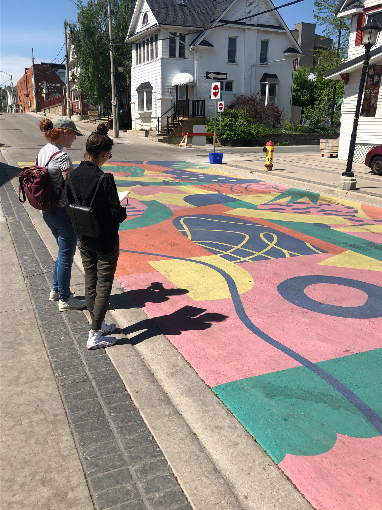 Two young women stand on a sidewalk looking down at a large colourful mural covering the road in a downtown Kitchener street
