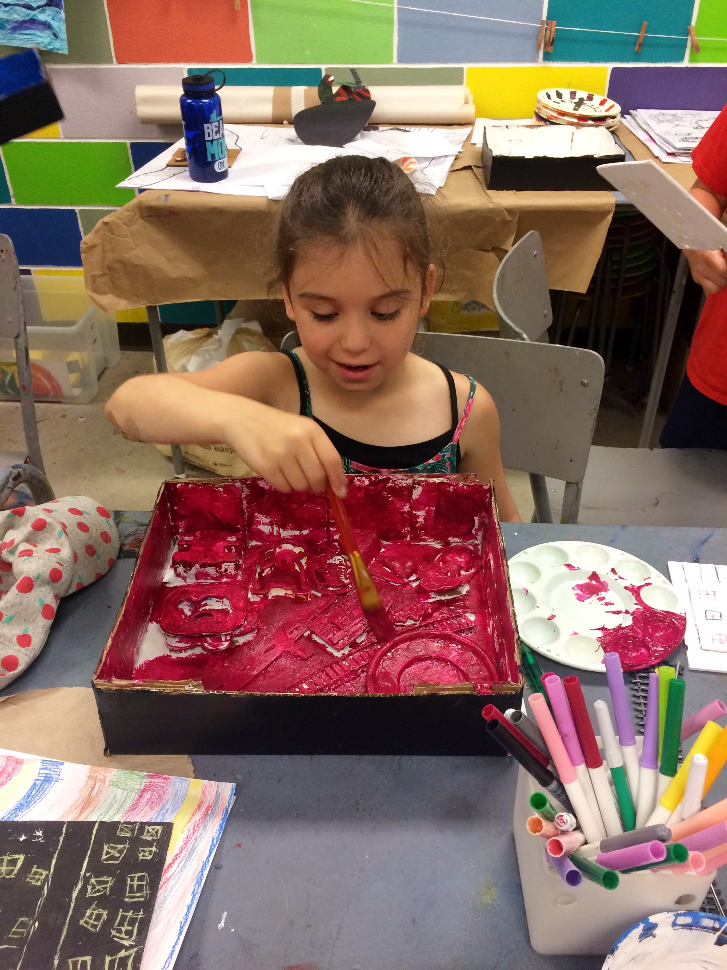 A young girl concentrates on applying bright magenta paint to the inside of a box filled with textured materials 