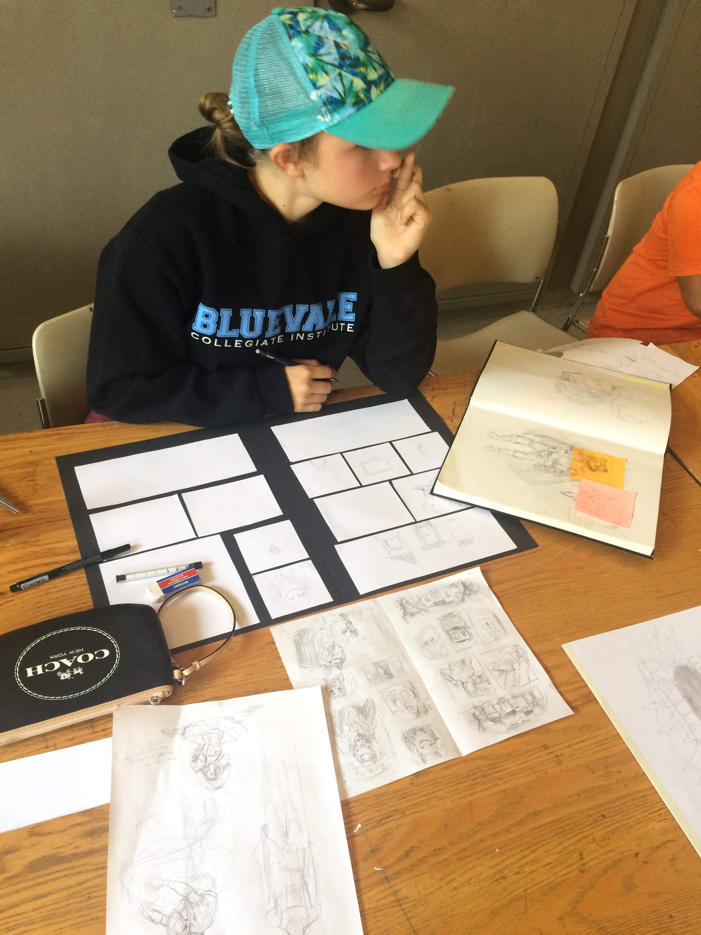 A young girl in a black sweatshirt and teal baseball cap sits at a desk before a series of sketches, notebooks and storyboards