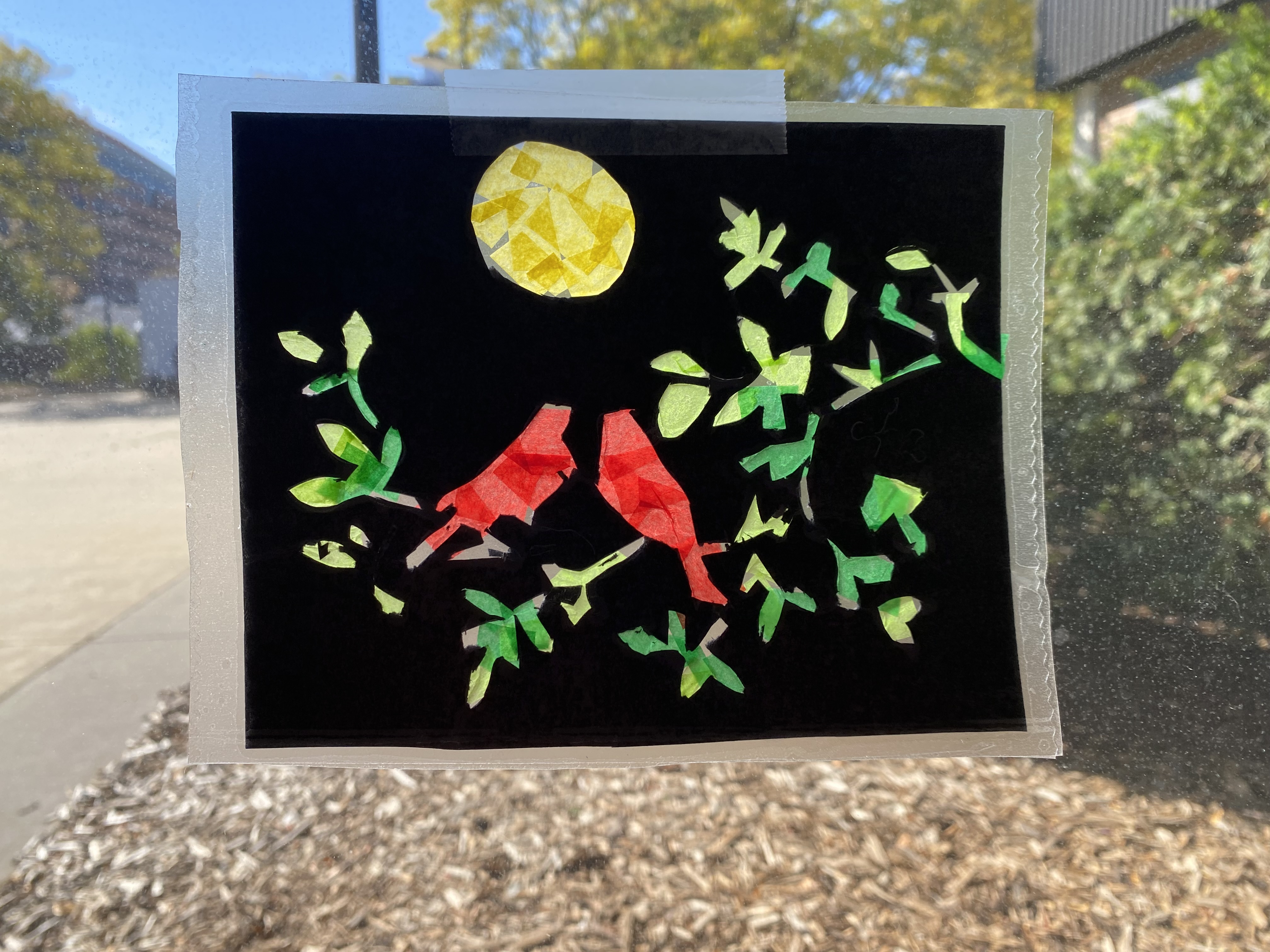 A tissue paper artwork of two red birds among green foliage below a yellow moon against a black background is taped to a window to allow light to pass through its shapes
