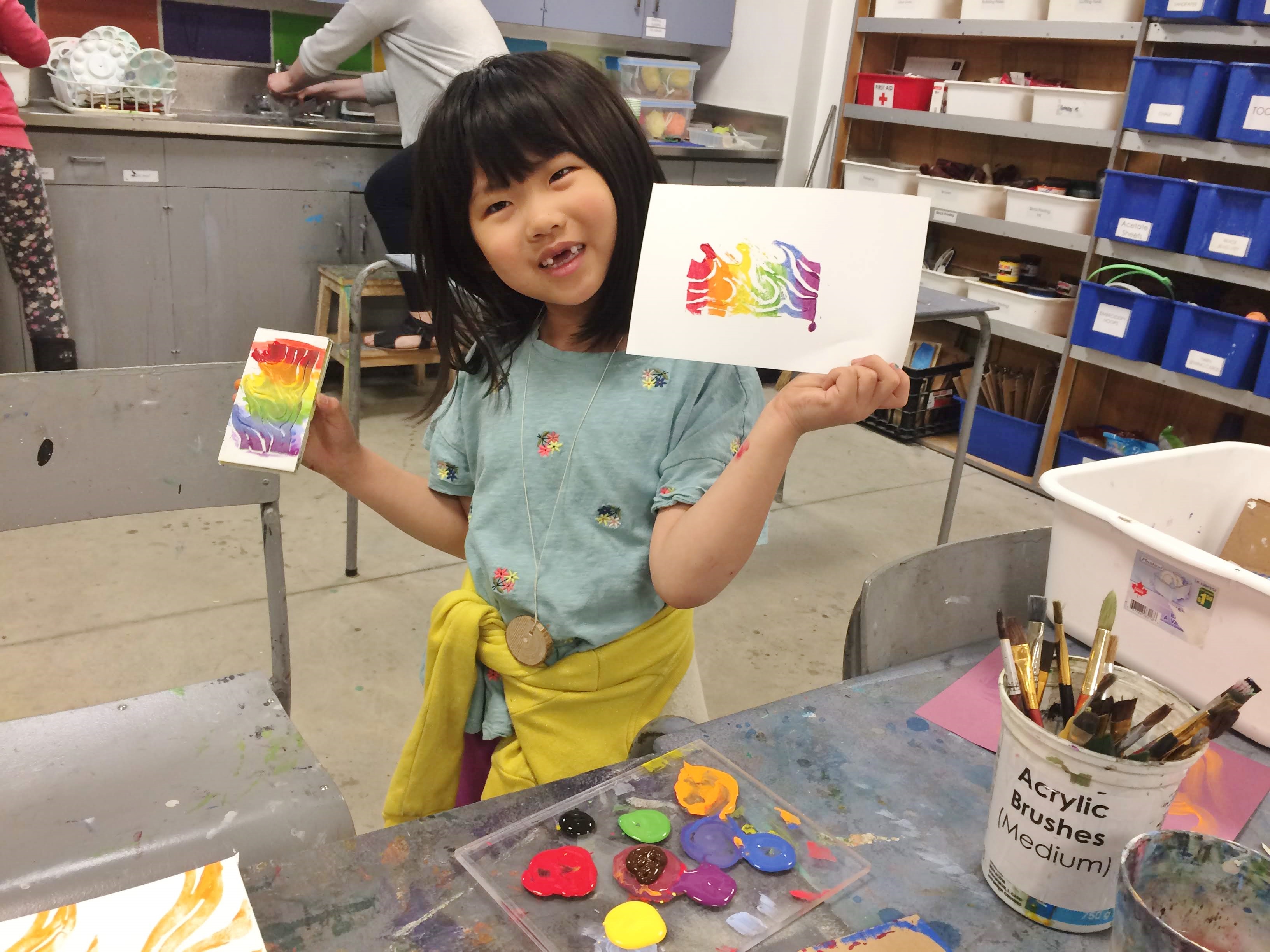 A young girl smiling as she holds up a rainbow print and her inked block in the KWAG studio