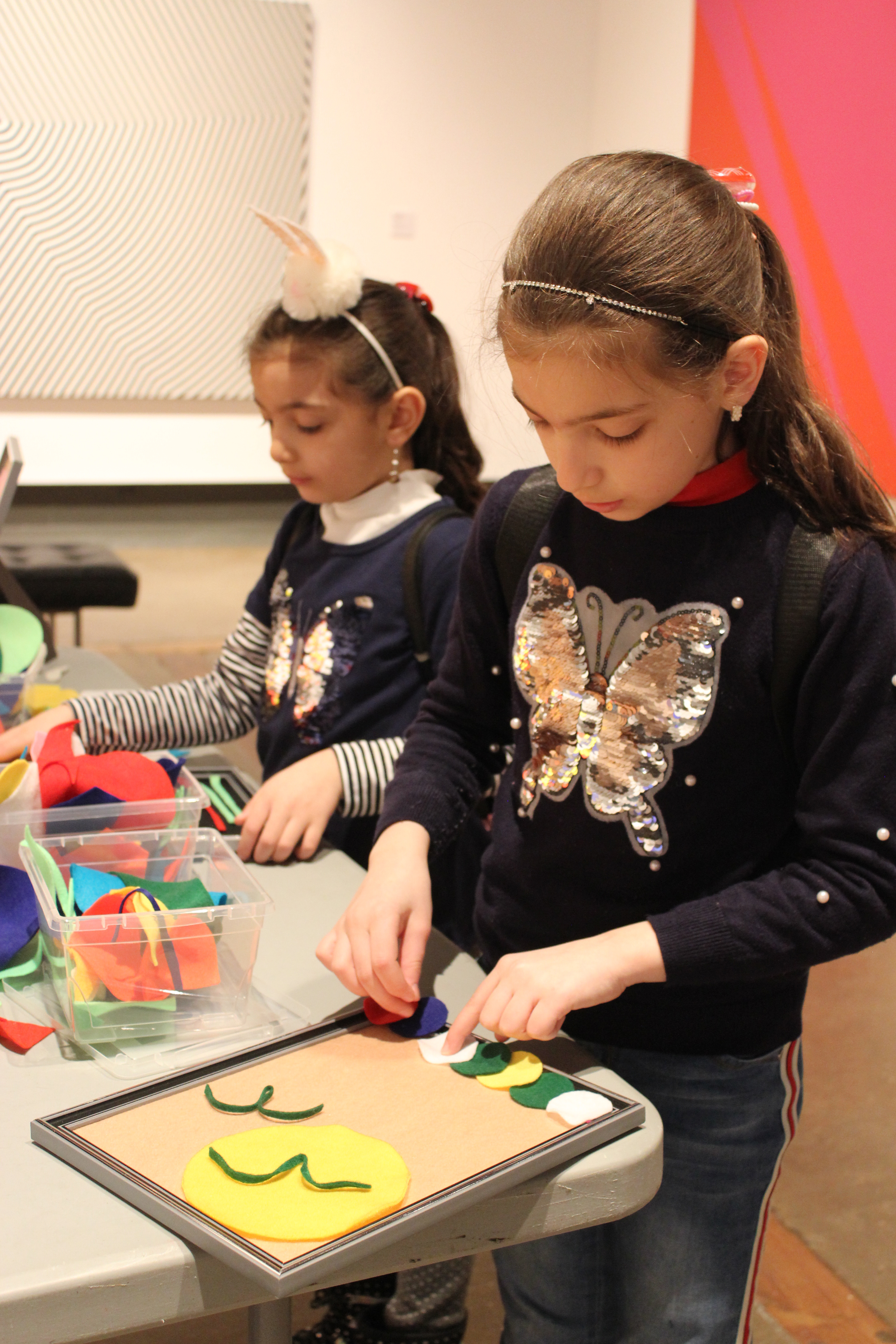 Two young girls arrange felt shapes into picture frames at a table set up in the Gallery