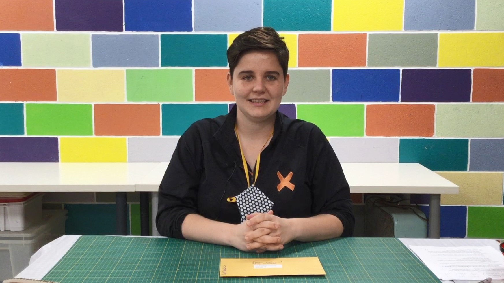 Photo of Brenda Reid, a smiling young woman with short-cropped brown hair wearing a black top, seated at a studio table in front of a brightly-painted brick wall