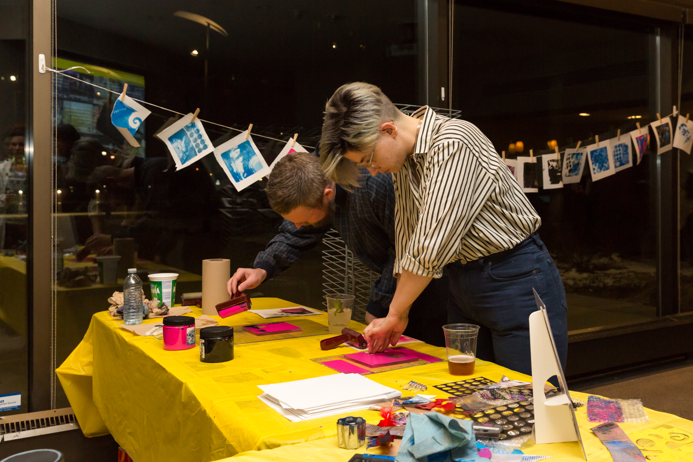 Two young people working on relief prints at a brightly-covered work table covered in printmaking supplies, alongside two cups of beer. A string of finished prints is hanging across a dark reflective window in the background