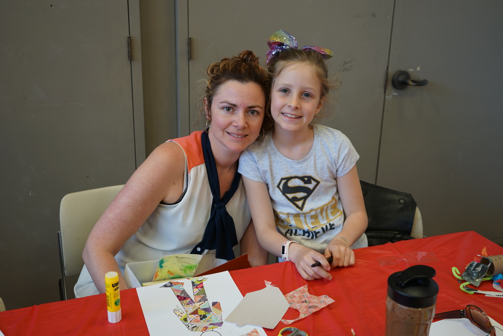 A mother and her daughter sit at table and smile at the camera with their artwork in front of them on the table