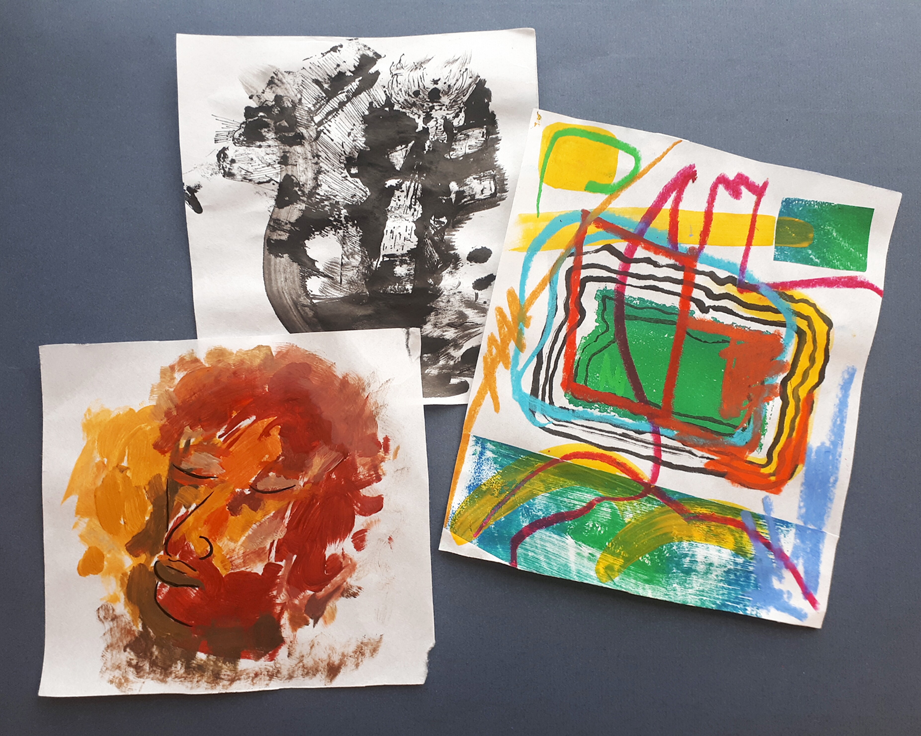 A set of three abstract paintings on paper arranged casually on a dark table surface