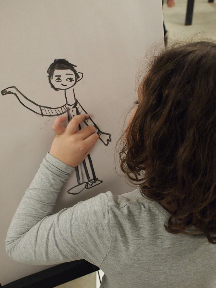 A young girl seen close-up from behind as she draws a posing figure on an easel pad with bold black lines