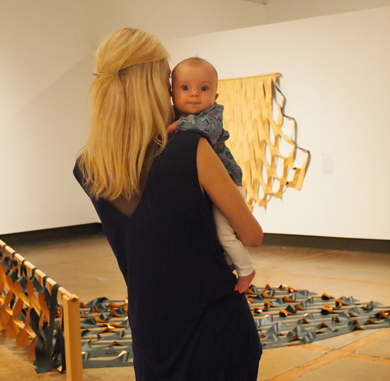 A young blonde woman with her back to the camera holding an infant who gazes over her shoulder towards the camera